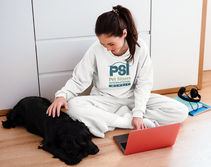 Adult woman with dark hair wearing a PSI member sweatshirt pets a dog by her side and looks on laptop