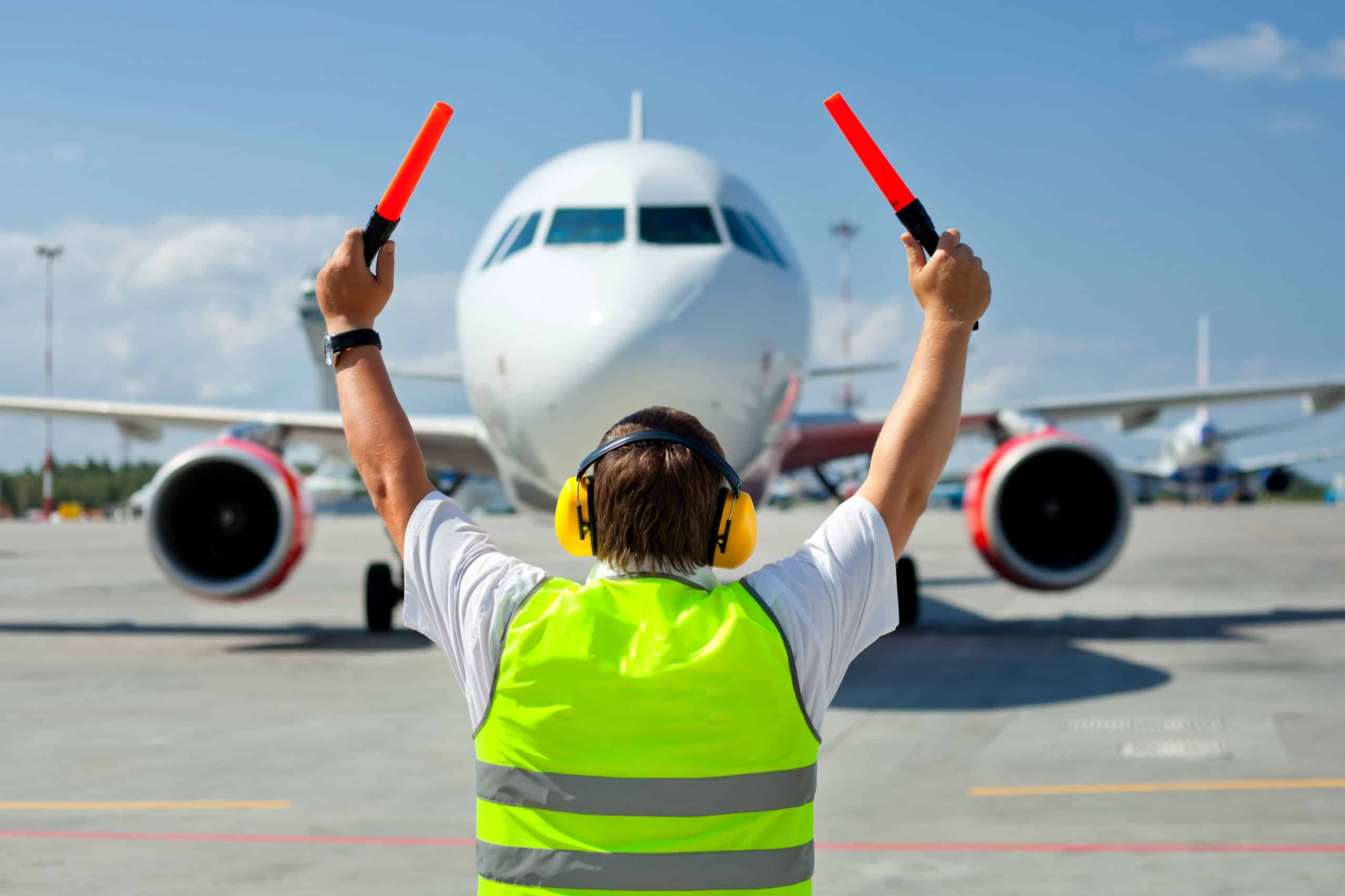 A ground crew worker in a reflective vest and ear protection guides an approaching airplane on the tarmac using two red signal batons.