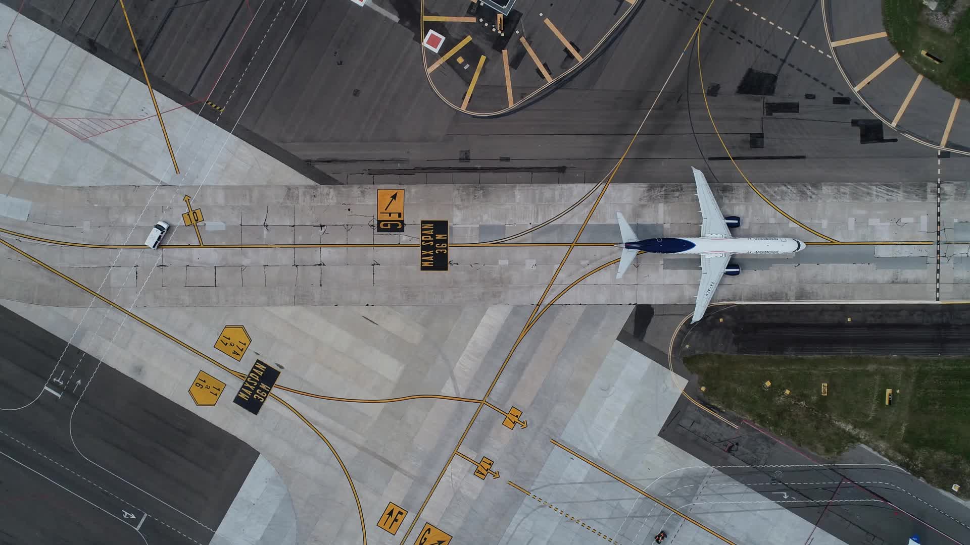 Aerial view of an airplane taxiing on an airport runway, with yellow directional markings and a small vehicle nearby. The runway and surrounding pavement are visible.