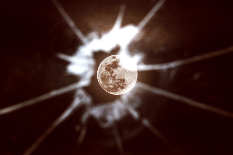 A close-up view of the full Moon with a bright, irregular white light pattern radiating outward against a dark background, creating a dramatic and surreal effect.