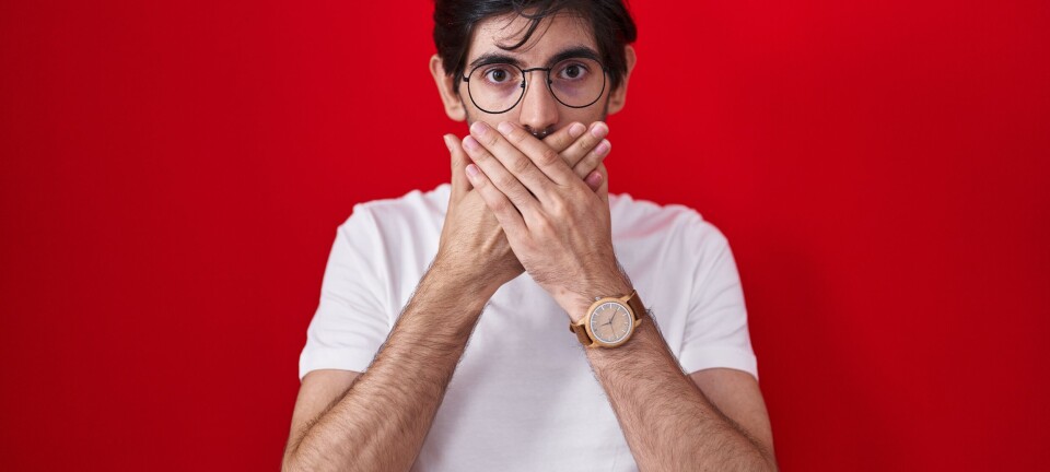 Young man in glasses covers his mouth with both hands against a red background.