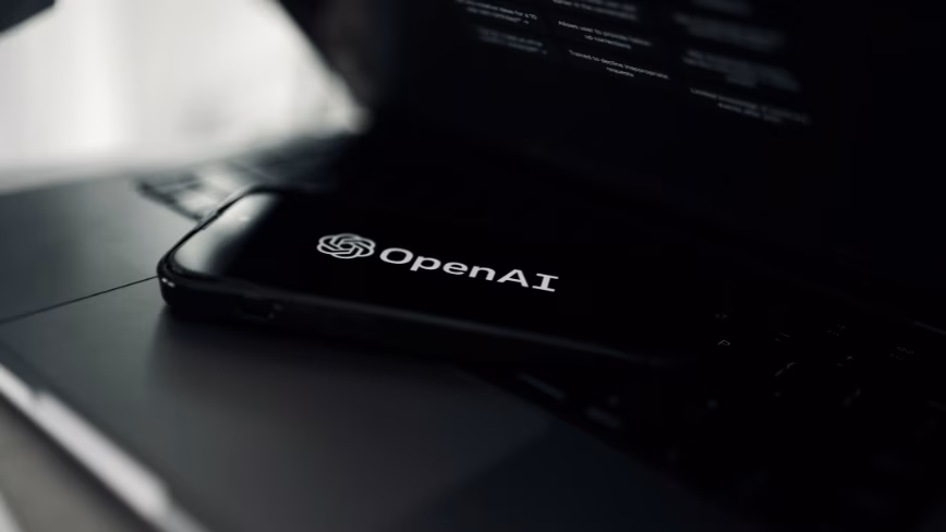A smartphone showing the OpenAI logo on a black screen, lying on a dark laptop keyboard, with a blurred chat interface on the laptop screen behind it.