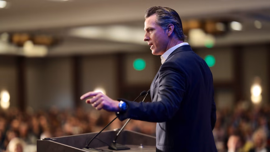 Gavin Newsom speaking at a lectern, gesturing towards an audience in a conference hall. Featured Image